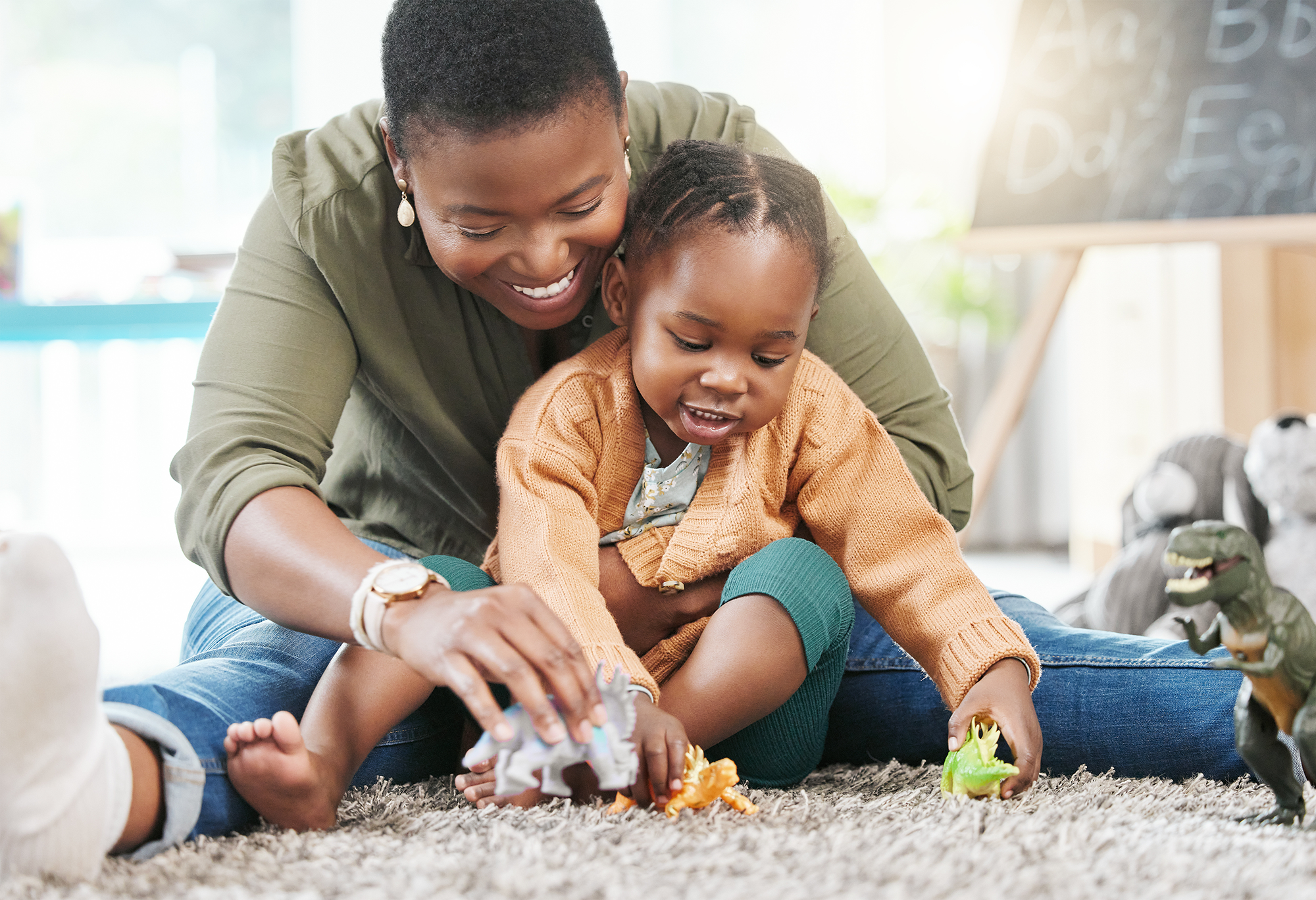 A woman and child sitting on a carpet, playing with toy dinosaurs.